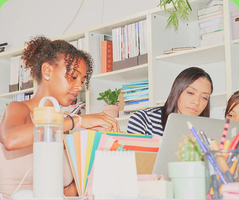 Two colleagues working at a desk in front of a colourful bookshelf — flat
