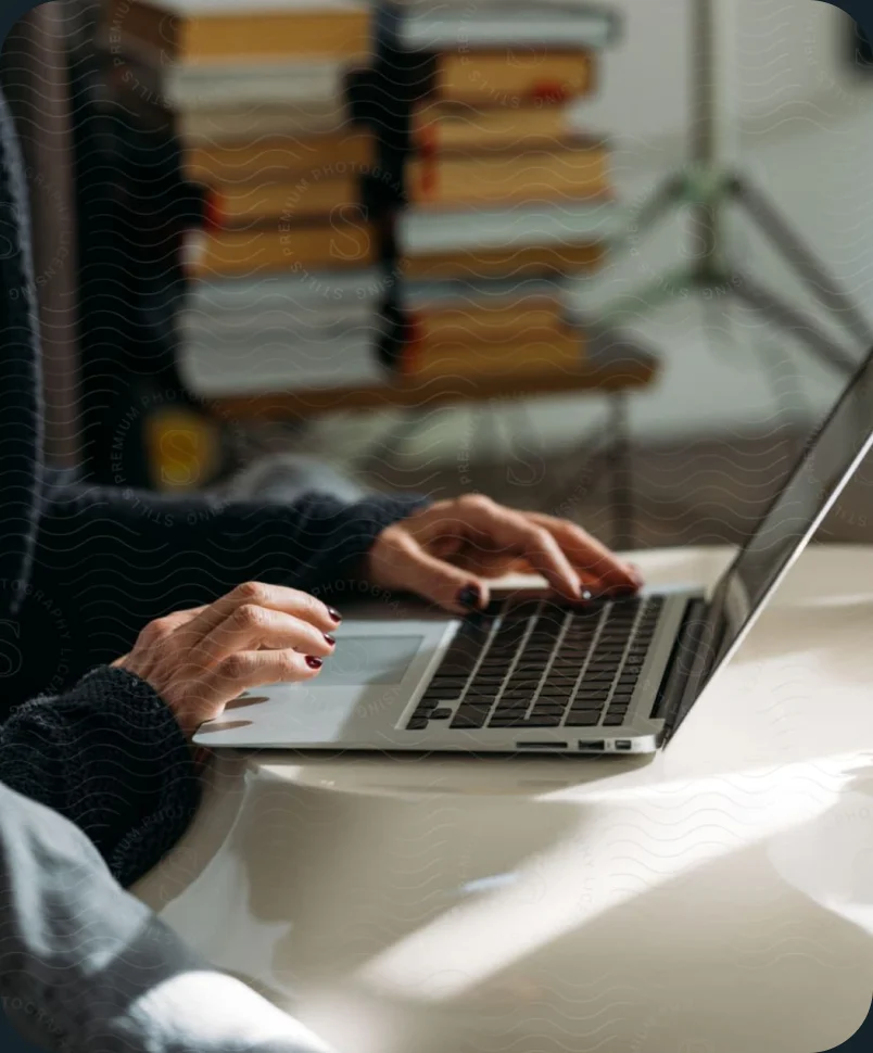 Close crop of a person typing on a laptop beside a stack of books