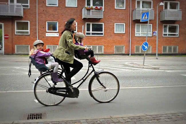 Courtesy of Comrade King A mother riding a bicycle, with two small children on attached seats.
