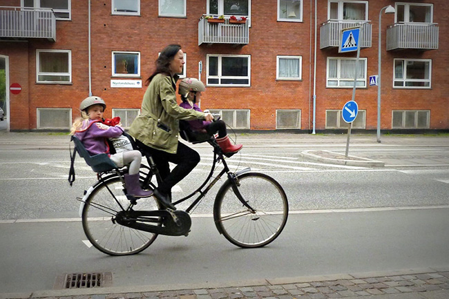A mother riding a bicycle, with two small children on attached seats. 