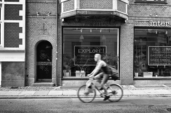 Courtesy of Sigfrid Lundberg Man riding a bicycle down a city street, past a sign that reads, “EXPLORE!”